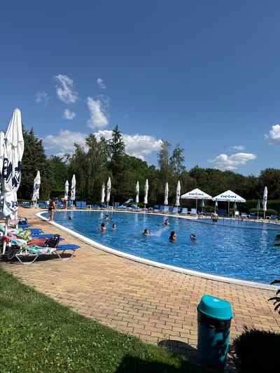 This is a horizontal, eye-level shot of an outdoor swimming pool area in Yagoda, Bulgaria. The bright blue swimming pool takes up a significant portion of the frame, with several people swimming or wading in it. The pool is surrounded by a tiled walkway in shades of yellow and red, and beyond that, a strip of green grass. A line of lounge chairs and white umbrellas with blue accents are positioned along the edge of the pool. Lush green trees and foliage form a backdrop behind the pool area. The sky is a clear, vibrant blue with scattered white clouds. The scene appears to be bathed in bright sunlight, suggesting it is a warm, sunny day, likely midday. The overall atmosphere is one of leisure and relaxation.
Visible text includes the word "DEVIN" repeated on several of the umbrellas. A large, teal-colored trash can with a black liner is also visible near the edge of the pool. The presence of lounge chairs, umbrellas, and people enjoying the water indicates a recreational setting, likely a public or resort pool.