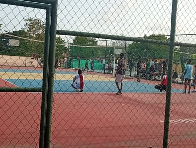The image shows a scene at a volleyball court in Ijanikin, Nigeria. The court is enclosed by a chain-link fence. The ground of the court is painted with red, blue, and yellow sections. Several people are present on the court and in the surrounding area. Some individuals are standing, while others are crouching or sitting. A volleyball net stretches across the center of the court, and a volleyball game appears to be in progress. Trees and buildings are visible in the background. The image is taken through the chain-link fence.