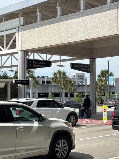 The image captures an airport scene, likely the Charleston International Airport in North Charleston, United States of America. A pedestrian is visible with their luggage near a crosswalk, indicated by a "STATE LAW STOP FOR CROSSWALK" sign. The setting is characterized by airport infrastructure including the canopy and signage "Courtesy/Public Transit → Ride Share", "Taxi/Shuttle Charter/Limo", "Shuttles / Taxis", and Zone 1. Several cars, including white sedans and a black SUV, are visible. Palm trees punctuate the urban landscape.