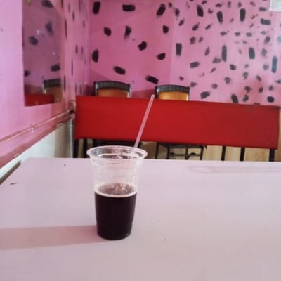 The image depicts a plastic cup filled with a dark liquid, likely soda or a similar beverage, with a straw inserted. The cup is positioned on a white table. Behind the table is a red bench or seat, and in the background, the wall is painted pink with irregular black blotches. The scene appears to be inside a commercial establishment, potentially a cafe or restaurant in Kinoo ward, Kenya. There are no people visible in the image.