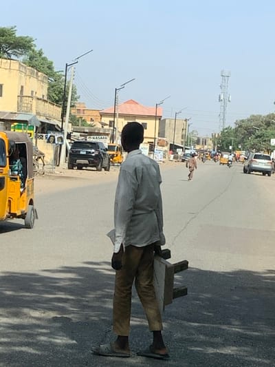The scene depicts an urban street in Hotoro South, Nigeria, under clear daylight. In the immediate foreground, facing away from the viewer, stands a male individual, appearing youthful, dressed in a light-colored long-sleeved shirt, brown trousers, and sandals. He carries a light-colored, possibly wooden, stool-like object under his left arm and holds a folded white item in his right hand.
The asphalt road, exhibiting visible cracks and repairs, features the main subject and, further down the road, another male figure in brown traditional attire walking in the center of the lane, also facing away. Various vehicles populate the street: a yellow auto-rickshaw (Keke Napep) with occupants is positioned on the left, closer to the foreground. Further down the road, multiple auto-rickshaws, motorcycles, and several cars, including a black SUV (Range Rover) parked on the left, indicate active traffic.
Along the left side of the road, a series of buildings are visible, including a multi-story structure with a light-colored facade and various smaller commercial establishments. One shop displays a sign with "NASARA." Utility poles and modern streetlights with solar panels line the road. In the background, on the right side of the street, a tall metal lattice communication tower is prominent. The sky is clear, uniformly light blue, and devoid of clouds.