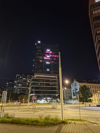 The image shows a nighttime view of the Spielberk Office Centre complex in Brno, Czechia. In the foreground, a wide road with multiple lanes is visible, with a pedestrian crossing marked. A white SUV is parked on the right side of the road. Several modern office buildings dominate the background. The central building, the Spielberk Office Centre, is illuminated from within with pink light. A tall, slender lamppost stands in the middle of the scene. Traffic lights are red, indicating that traffic is stopped. An older, ornate building with arched windows is visible to the right of the modern office complex. A large advertisement is displayed on the side of another building. The sky is dark, typical of a night scene. The overall impression is one of a modern urban environment at night.