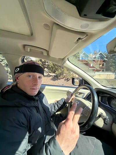 A male individual is seated in the driver's seat of a Volvo vehicle on a sunny day in La Salle-les-Alpes, France. He has light skin, blue eyes, and a neutral expression, looking directly at the viewer. He wears a black Oakley headband over a multicolored patterned headwrap, a silver hoop earring in his left ear, and a dark, possibly black or grey, puffer jacket. His right hand is positioned on the steering wheel, while his left hand is raised in front of the steering wheel, displaying an extended index and middle finger gesture. A small tattoo is visible on his left wrist. The vehicle's interior features a beige dashboard and ceiling, a sunroof, and a sun visor. The instrument cluster shows a numerical reading of "24" and "D" for the gear. Through the passenger-side window and windshield, a multi-story chalet-style house with light brown wooden siding and distinctive blue-grey gabled roofs is visible, partially obscured by pine trees. The background also reveals a clear blue sky and some dry ground patches.