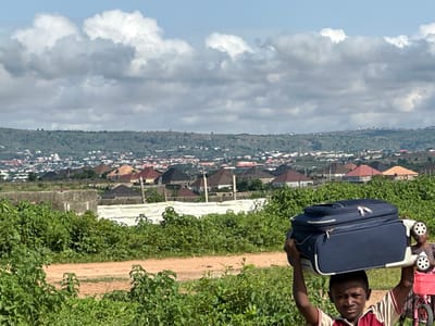 A young boy carries a large, dark blue suitcase on his head while standing in a grassy area with a dirt road in the foreground. Behind him, a younger child stands next to a pink bicycle with a basket. In the background, a sprawling city with red-tiled roofs stretches across a hilly landscape under a cloudy sky. This scene is located in Gidan Waziri, Nigeria.