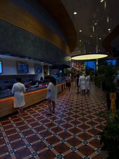 This image depicts an indoor food service area or bar within Therme Bucuresti, located in Săftica, Romania. The scene is illuminated by artificial lighting, indicating evening or nighttime, with large windows in the background revealing a dark sky.

In the foreground, two women stand at a long counter. The woman on the left, with her back to the viewer, wears a white bathrobe. The woman on the right, also facing away, wears a loose white tunic over dark shorts and flip-flops, holding a mobile phone. The counter features a light wood base with an orange LED strip illumination, a white subway-tiled backsplash, and glass display cases. Three digital menu screens displaying text and graphics are mounted above the counter. Behind the counter, a person in a light blue shirt is visible.

The floor is covered with patterned tiles featuring a geometric design in shades of terracotta, beige, and blue. In the mid-ground and background, several individuals are present; some wear white bathrobes, others are in swimwear (e.g., black swim shorts), suggesting a spa or aquatic leisure facility. A group of people in bathrobes stands conversing on the right.

The ceiling features a complex architectural design with curved panels of light wood and dark grey material, incorporating recessed lighting. A large, circular illuminated fixture is prominent in the background ceiling. Numerous large potted plants are strategically placed throughout the area, contributing to a lush, tropical ambiance. A colorful mural with large floral patterns is visible on a wall behind the plants and near the windows. A yellow "wet floor" caution sign is partially visible in the bottom right corner.