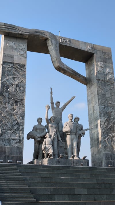 The image shows a concrete monument in Phan Rang–Tháp Chàm, Vietnam. The monument features a large archway with bas-relief carvings of battle scenes. Within the archway is a group of sculpted figures, possibly representing soldiers or civilians. To the right of the figures, there are gray stone steps. The sky visible through the arch is clear and blue. The date "16-4" is visible on a part of the archway.