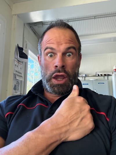 A medium shot shows a man with a beard and dark hair in a black shirt with red piping, making a surprised facial expression. He is indoors, near a window. Papers are visible on the windowsill. In the background are cabinets and a white, horizontally-lined ceiling. The location is not identifiable as Diekirch, Luxembourg, based on the image.