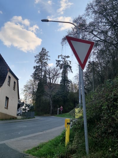 The image presents a daytime outdoor scene in Obernburg am Main, Germany. No individuals are present.
In the foreground, to the right, is a grassy embankment interspersed with dense green and brown foliage. A grey metallic pole rises from this embankment, supporting a prominent "Give Way" (yield) traffic sign, which is an inverted equilateral triangle with a thick red border and a white interior. Below this sign, a yellow short cylindrical bollard is visible in the grass. A yellow rectangular sign with black German text, partially obscured by greenery, is affixed to the sign pole, displaying fragmented words like "SUCH..." and "HAUS..." and a phone number "0151-611...". A street light fixture with a curved arm extends from the top of the pole.
A paved asphalt road curves gently to the right through the midground, with faint white markings on its surface and a light grey curb separating it from the grassy verge. To the left of the road, a two-story residential building with a light-colored (cream/beige) facade and a dark reddish-brown tiled gable roof is partially visible. It features multiple rectangular windows with brown frames. A silver passenger vehicle is partially visible parked behind a dark fence adjacent to the house. Further down the road on the left, a smaller triangular warning sign and a circular sign are visible on a pole.
The background is characterized by numerous trees, many appearing bare or sparsely leafed, suggesting a colder season. Dominating the central background is a church building with a dark roof and a distinct steeple, partially obscured by the surrounding trees. The sky above is light blue with scattered white cumulus clouds.