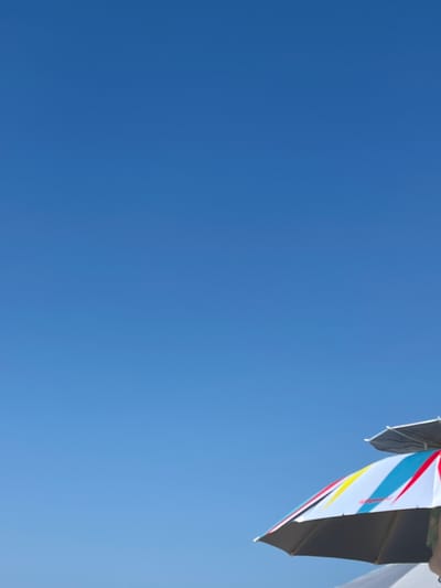 The image captures a bright blue sky, suggesting a clear and sunny day, likely in the daytime given the intensity of the blue. In the upper right corner, a portion of a structure, possibly a building or canopy, is visible. Attached to this structure are several colorful flags or banners. These fabric items are a dominant feature in this section of the image, with visible stripes of white, blue, red, and yellow. Some of the banners appear to be layered or folded, creating a dynamic visual. The rest of the frame is filled with the uninterrupted expanse of the sky, indicating an outdoor setting with a wide-open view. There is no visible text in the image. The overall impression is one of a bright, open, and possibly festive atmosphere, given the presence of colorful flags against a clear blue sky. The location is identified as Ahtopol, Bulgaria, which is a coastal town, and the clear sky and vibrant colors could be indicative of pleasant weather often experienced in such locations.