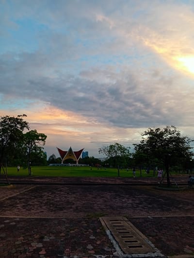 The image depicts an outdoor scene at dusk in Bang Kaeo Subdistrict, Thailand. In the foreground, a paved area made of reddish-brown interlocking bricks is visible, with a concrete drain cover in the lower part of the frame. Beyond the paved area is a large, manicured green lawn. In the middle ground, a distinctive red and yellow open-air structure, possibly a stage or pavilion, is situated. Several trees of varying sizes are scattered throughout the scene, with foliage and some exposed branches. In the background, buildings and the sky with scattered clouds are visible. The sky displays soft hues of orange and yellow, indicative of sunset or twilight. Several individuals are present on the lawn, engaged in various activities such as walking, standing, or possibly playing. The overall atmosphere suggests a public park or recreational area.