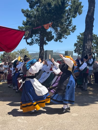 In the city of Llanera, Spain, a group of people are participating in a cultural festival. The scene captures a lively moment as several individuals, adorned in traditional folk costumes, engage in a dance. The costumes are vibrant, featuring colorful skirts, blouses, and headscarves. One dancer wears a distinctive blue headscarf, while others have yellow and white coverings. The dancers are captured in motion, with their arms raised and skirts swirling.
To the right, a bagpiper plays a traditional instrument, his bagpipe clearly visible. Beside him, a drummer beats on a drum, adding to the rhythmic atmosphere of the event. In the background, other festival-goers observe the performance, some standing and watching, while others are partially obscured. The environment suggests an outdoor gathering, with a large pine tree dominating the upper right portion of the frame. Red and orange banners are strung overhead, contributing to the festive ambiance. The ground is a mixture of dirt and paved surfaces, indicating a well-established event space. The sky is clear and blue, suggesting a sunny day. The overall impression is one of a vibrant cultural celebration in Llanera, Spain.
