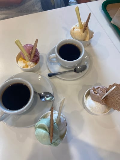 A high-angle shot captures a tabletop scene featuring four small bowls of gelato and two cups of black coffee, along with spoons and thin, rectangular wafers. The gelato bowls hold different flavor combinations. From the top left going clockwise, there is a bowl with what appears to be light yellow gelato, then a bowl with chocolate and vanilla gelato topped with a large, round wafer, a bowl containing green and white gelato, and lastly a bowl with purple and yellow gelato. All the bowls are paired with a spoon. Two coffee cups, each on a saucer, are placed near the bowls of gelato. The scene takes place on a light colored table, but there is not enough context to determine the location in Lecco, Italy.