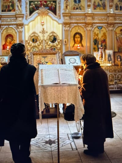 Two individuals, facing away from the camera, stand in an Orthodox church in Pastavy, Belarus. The person on the left wears a dark hooded garment and a dark beanie. The person on the right wears a dark coat and a brown knit hat. Both are positioned behind a stand displaying an open book, adorned with a lace cloth. A lit candle is visible near the person on the right. The background features religious icons and ornate architectural details typical of an Orthodox church interior. The floor is tiled with a geometric pattern.