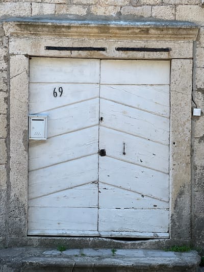 The image presents a static, head-on view of an aged, white double-leaf wooden door, designated by the house number "69", situated within a weathered stone architectural frame in Bogdašići, Montenegro. The door's white paint is extensively chipped and peeled, revealing the underlying material and indicating significant exposure. Each door leaf is composed of multiple horizontal planks, arranged to create a chevron or diagonal pattern across the central sections. A small, white, rectangular metal mailbox with lower perforations is affixed to the upper left door leaf, positioned below the "69". The right door leaf features a dark, prominent latch or lock mechanism, with a corresponding dark pull handle on the left leaf. Two recessed black horizontal metal bars are set into the stone lintel directly above the door. The surrounding wall is constructed from irregularly shaped, light-colored stone blocks. To the right of the door frame, a small, white rectangular electronic device, likely a doorbell or intercom component, is mounted. A worn stone step or threshold forms the base of the entrance, with sparse green vegetation emerging from cracks in the stone. The scene is illuminated by direct sunlight, highlighting textures and casting subtle shadows.