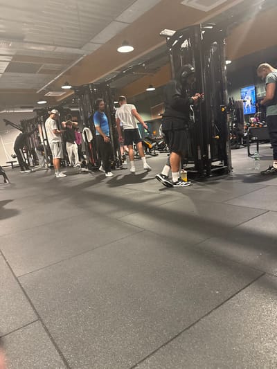 The image shows a gym setting with multiple individuals using cable machine equipment. They are lined up in a row, facing the machines, engaged in various exercises. Most are wearing athletic attire, including shorts, t-shirts, and athletic shoes. A television screen can be seen in the background. The gym has a gray tiled floor and a modern, well-lit environment.