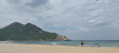 The image depicts a coastal scene in Cát Tiến, Vietnam. A wide sandy beach stretches across the foreground. In the background, a large, verdant mountain dominates the left side of the frame, its slopes covered in dense vegetation. The sea, a clear turquoise, meets the sandy shore, with gentle waves lapping at the beach. Several people are visible in the water and on the beach. On the far right, a man stands facing away from the camera, holding a bag. Closer to the water's edge, a group of people are gathered. Scattered across the turquoise sea are numerous small boats. The sky is overcast with a mix of white and grey clouds.