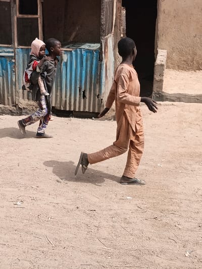 The image depicts three individuals in an arid, unpaved area of Zango, Nigeria, under bright sunlight.

In the foreground, a child, likely male, walks from left to right. This child wears a light brown or tan long-sleeved tunic with matching trousers and dark flip-flops. Their right foot is lifted mid-stride, and both arms are slightly outstretched from the body with open hands, palms down, fingers spread, while looking towards the right and slightly upward.

To the left and slightly behind, another child, also appearing male, walks in the same direction, carrying a younger child piggyback. The older child wears a dark grey long-sleeved top and patterned trousers with blue, white, red, and black motifs, with light grey slip-on footwear. The younger child on their back wears a light pink bonnet and a red, white, and black patterned garment, looking to the right.

The ground is uniformly light brown, dry earth with scattered small debris. In the background on the left, a structure with a distressed, light blue corrugated metal wall and an upper window is visible. To the right, a weathered light brown wall with an open, dark doorway is present. Shadows cast on the ground indicate direct overhead sunlight.