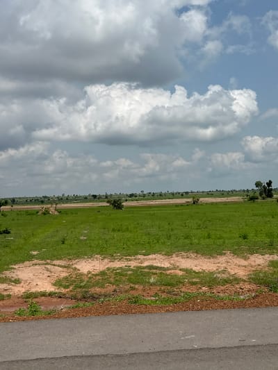 The image depicts a vast, open expanse of grassland under a cloudy sky. The foreground features a paved road with a border of reddish-brown dirt and scattered vegetation. Beyond the road, the terrain is covered in lush green grass, with patches of exposed reddish-brown earth. In the middle ground, the landscape continues with more greenery and scattered trees. A thin line of trees marks the distant horizon, beyond which the sky is filled with a mix of white and gray clouds against a pale blue backdrop. The scene is devoid of any people or human activity, suggesting a rural or undeveloped area. The location is identified as Serakye, Nigeria.