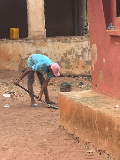 In Aguata, Nigeria, a lone male individual is depicted outdoors, bent forward at the waist. He wears a light blue t-shirt, stained with what appears to be dirt or mud, dark knee-length shorts, and blue and white flip-flops. A pink baseball cap covers his head. His arms are extended downwards, holding a dark-colored shovel with both hands, its blade positioned on the reddish-brown dirt ground. A red wristband is visible on his left wrist.

The immediate environment consists of loose, reddish-brown soil, marked with small indentations and scattered pieces of white and colored litter. To the right, a solid red-orange concrete structure, possibly a low wall or bench, extends into the foreground. In the mid-ground, behind the individual, a light gray bucket with dark banding is visible on the ground, next to additional scattered debris and sparse vegetation. Further behind, partially obscured, are sections of weathered, light-colored concrete or stucco walls belonging to a larger, unfinished or deteriorated building. This structure features open window or door frames, and a prominent concrete pillar. A yellow plastic jerrycan rests near one of the building's walls in the background. The scene is illuminated by natural light, casting no distinct long shadows, suggesting an overhead light source.
