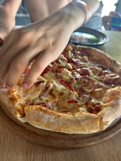 A close-up shot shows a person's hand reaching for a pizza. The pizza is golden brown with melted cheese and red pepper slices visible on top. The crust is thick and appears to be cooked to perfection. The pizza is resting on a wooden board, which is placed on a wooden table. In the background, another person is partially visible, wearing a blue shirt. The lighting suggests it might be daytime, possibly outdoors or in a well-lit room. The scene evokes a sense of casual dining and enjoyment. The focus is on the pizza and the hand, highlighting the act of taking a slice.