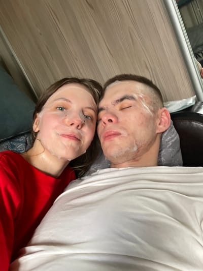 Two individuals are the primary subjects of the image. The person on the left has a white facial mask on and is wearing a red t-shirt. The person on the right has a similar white substance on their face and is wearing a grey t-shirt. A white garment is draped over the right individual. A dark leather surface is visible in the upper-right corner of the frame. The background includes a light wooden surface. The photograph's location is speculated to be within a domestic environment, potentially Ulukulevo, Russia, based on the prompt's specification, but there is no evidence of the location in the image.