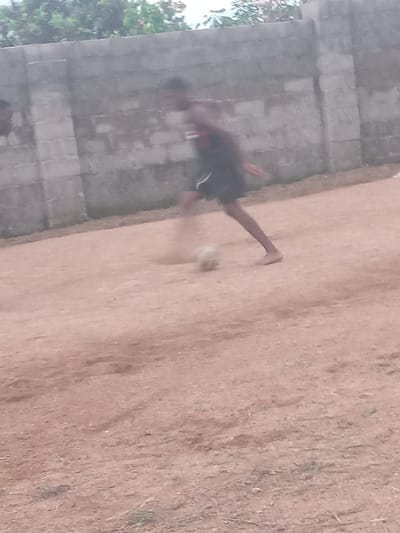 A barefoot person in dark shorts is kicking a light-colored ball on a dirt surface in Achara Ndiagu, Nigeria. A concrete block wall is visible in the background, along with some trees. The photo is blurred, indicating motion.