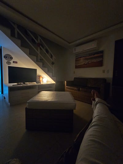 The image depicts the interior of a dimly lit home, possibly a living room, captured from a low angle. On the right, a person is seen sitting or lying on a sofa, with their legs and lower torso, clad in light-colored pants, visible. The sofa appears to be made of a dark, woven material, possibly wicker, and an ottoman or bench with a light top and dark base is in the foreground.

To the left, a white-railed staircase ascends, with a small landing area beneath it that houses a television screen, a lit lamp, and various decorative items. Circular emblems or plaques, some with visible text like "1940" and "WHISKIES", adorn the wall near the stairs. Above the landing, a white air conditioning unit is mounted on the wall, and a framed piece of abstract or landscape art hangs above it.

The overall scene suggests an evening or nighttime setting due to the reliance on artificial lighting, primarily from the lamp under the stairs and some ambient light. The furnishings and decor indicate a modern residential space. The person appears to be in a state of relaxation. While the location context is Cabo Frio, Brazil, there are no specific visual cues within the image to confirm this.