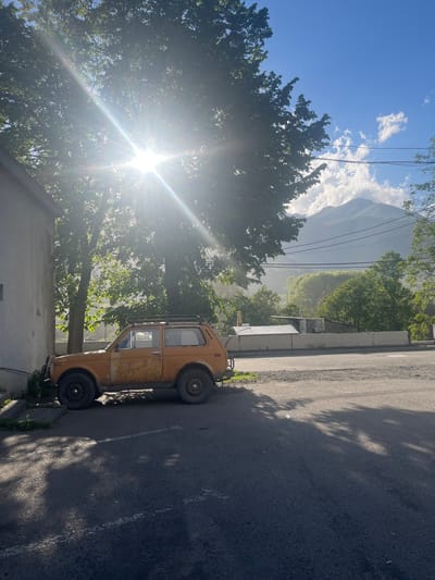A bright sun shines through the leaves of a large tree, casting dappled shadows on the ground and an orange, old-fashioned utility vehicle parked on an asphalt road in Stepantsminda, Georgia. The vehicle, appearing weathered and rusty, is positioned to the left of the frame, adjacent to a pale, textured building. Behind the vehicle and across the road, a low concrete barrier encloses several buildings and greenery. In the background, a large, misty mountain range dominates the horizon, under a bright blue sky dotted with white clouds. Electrical wires are visible strung across the sky. The scene appears to be a street view in the town of Stepantsminda.
