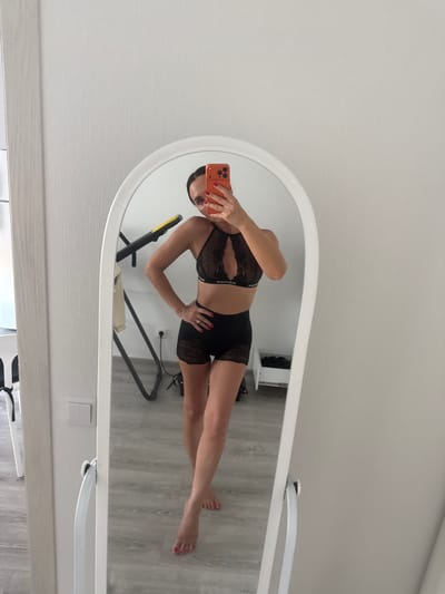A woman is taking a mirror selfie in a room. She is wearing black lace lingerie, consisting of a bralette and shorts. She is holding an orange smartphone to take the photo. Her left hand is on her hip and her right arm is extended to hold the phone. The room has light-colored walls and a light wood-grain floor. In the reflection of the mirror, a piece of exercise equipment and a white drawer unit can be seen. The image was taken in Zapadny, Russia.