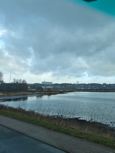 The photograph depicts a body of water in Kolding, Denmark. The sky is overcast, with a prominent cloud cover. In the background, there are buildings, trees, and a crane visible along the distant shoreline. Several sailboats are anchored in the water near the shoreline. The foreground consists of a waterfront area with grass and a paved walkway. The photograph appears to be taken from an elevated position, possibly from inside a vehicle.