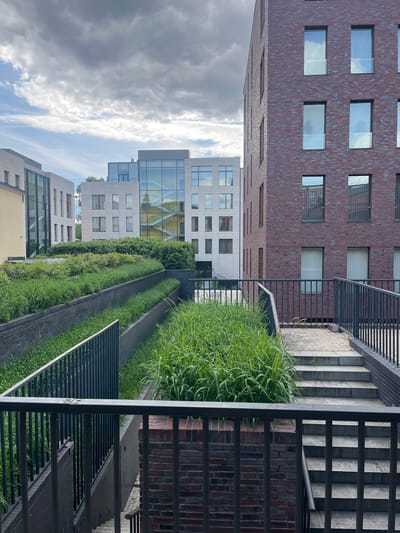 The image depicts a vertically-oriented urban scene with modern architecture under a partly cloudy sky. The primary subjects include multiple buildings with varying facades. One building is constructed of reddish-brown brick with evenly spaced windows, while others are clad in light-colored materials with glass panels. There is a vertical green space or garden visible between the buildings, featuring linear sections of grass and vegetation, as well as a round bush. A metal staircase with railings is present on the right side of the image. The location is identified as Перовское сельское поселение, Ukraine. There are no people visible in the image. The environment includes a mix of natural and constructed elements.