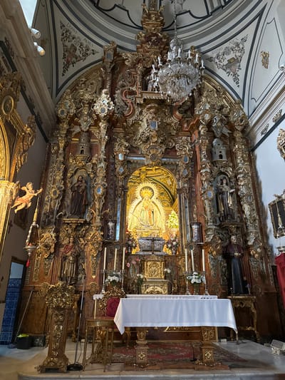 The image displays the ornate Baroque main altarpiece of the Church of Santa María la Mayor in Ronda, Spain. The altarpiece is a multi-tiered, gilded wooden structure featuring extensive gold leaf ornamentation over a dark wooden base.

At the center, within a brightly lit, deeply recessed golden niche, is a statue of the Virgin Mary, likely the Virgen de la Paz, adorned with a radiant golden halo and rich vestments. Below this central figure, on the altar table, a smaller golden tabernacle or reliquary is positioned, flanked by a silver vessel and two tall white candles in ornate gilded holders. Two decorative vases containing white and pink floral arrangements are also present on the altar. The altar table is covered with a white linen cloth, and its base is gilded. A patterned red and beige rug lies on the floor in front of the altar.

The altarpiece incorporates multiple statues: a male saint holding a child (possibly Saint Joseph with the Infant Jesus) in the upper left, a female saint holding a book or staff next to a small tower-like structure in the upper right, a robed male saint in the lower left, and a seated ecclesiastical figure, resembling a Pope or Bishop, in the lower right.

To the left of the altar, an elaborate gilded lectern with a microphone stands. An angelic figure in flight, also gilded, extends from a side architectural element on the left wall, above a section of blue and white tiled wainscoting. The vaulted ceiling above is adorned with frescoes featuring striped patterns, floral motifs, and Latin inscriptions, including "DOMVS AVREA" (House of Gold) on the left and "FOEDERIS ARCA" (Ark of the Covenant) on the right. A large, multi-tiered crystal chandelier hangs centrally from the ceiling in front of the altarpiece. No individuals are present in the scene.