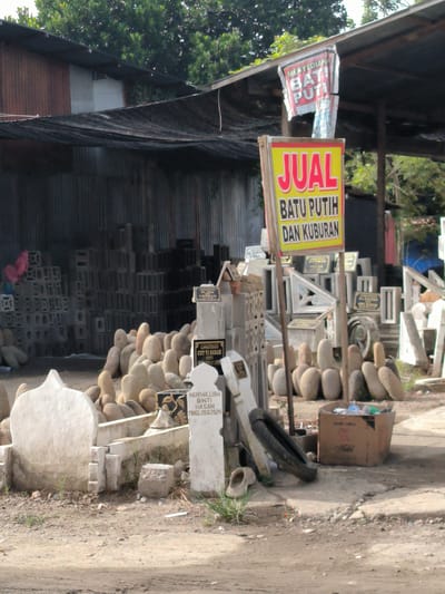 This is a medium shot of a street-side stall in Lhoknga, Indonesia, specializing in tombstone sales. The foreground is dominated by a white, dome-shaped tombstone on the left and a rectangular tombstone with text and decorative elements on the right. Several smaller, smooth, light-brown, oval-shaped stones are piled behind these. A wooden sign with red text on a yellow background, advertising "JUAL BATU PUTIH DAN KUBURAN" (White Stone and Graves for Sale), stands prominently in the center. Behind the tombstones, stacks of gray concrete blocks are visible, some arranged in intricate patterns. A black mesh tarp is stretched over a wooden frame, providing shade. In the background, trees with dense green foliage can be seen. A brown cardboard box filled with plastic bottles and other debris sits near the right side of the frame. The ground is a mixture of dirt and gravel.