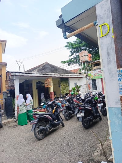A street scene features parked motorbikes lining the side of a residential building. Two women in matching headscarves and green skirts stand near the bikes. A water tower is visible behind the building, and the "D" of a sign is seen in the foreground. A utility pole is in the background and a building facade. The road surface is asphalt.