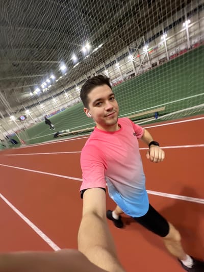 A man poses for a selfie on an indoor track. He wears a pink and blue shirt, black compression socks, and running shoes. The track is red with white lane markers. A green netted area with stadium seating is visible in the background. The building has a high ceiling with industrial lighting. The man is running mid stride.