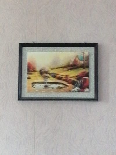 The image shows a framed painting hanging on a textured light-colored wall. The painting depicts a landscape with a house, trees, and a pond. The style appears to be impressionistic, with visible brushstrokes and a soft, blended color palette. The house in the painting has a light blue roof and a red-brown body. A path or walkway curves around the pond, and there are areas of green grass and colorful foliage, suggesting a garden or park setting. The overall impression is one of a serene, rural scene. The painting is framed by a thin black border. The wall behind the painting has a subtle, repeating pattern.