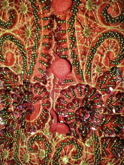 This image presents a forensic, close-up view of an elaborately decorated textile. The base material is a vibrant reddish-orange fabric, covered extensively with intricate embroidery executed in a shimmering golden-yellow thread. This embroidery forms complex curvilinear patterns, stylized floral motifs, and interconnected designs across the surface. Embedded within and alongside these embroidered patterns are numerous small, olive-green seed beads, arranged in dense lines and clusters, providing additional texture and outlining. Scattered throughout are clusters of iridescent sequins, predominantly red and purple, which reflect light and are sewn onto the fabric in overlapping formations, creating a glittering effect. Several small, spherical or tapered elements, constructed from tightly wrapped or stitched red fabric, are also affixed to the surface. The scene exclusively depicts this decorated fabric; no people, actions, interactions, or wider environmental context are visible. The image does not visually depict any specific location within the city of Keo Kouang, Laos, or any other geographical setting.