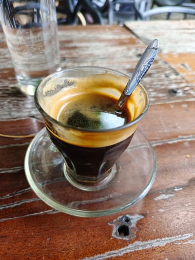 A close-up, eye-level shot shows a glass of dark coffee with a thick layer of crema, resting on a glass saucer on a weathered wooden table. A spoon with a decorative pattern is partially submerged in the coffee. To the left of the coffee cup, a partially filled glass of water is visible. The background is blurred, showing glimpses of chairs and possibly a bicycle, suggesting an outdoor or semi-outdoor seating area, likely at a cafe or warung in Pantegajah, Indonesia. The lighting suggests it is daytime.