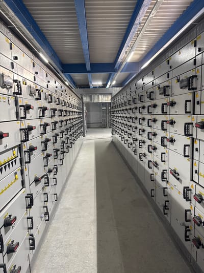This is a medium-shot, eye-level, vertical photograph of a long electrical panel room. The room is filled with rows of numerous grey electrical panels, each with black handles and red switches. The panels are arranged in a grid system and extend far into the distance, creating a sense of depth and scale. The lighting appears to be artificial and is evenly distributed throughout the space, casting minimal shadows.

The scene is industrial and appears to be in a large, enclosed facility, possibly a substation or a power distribution center. The walls are a textured grey concrete, and the ceiling is not fully visible but appears to be of industrial construction. There is a blue structural beam running along the left side of the image, supporting a corrugated metal wall or ceiling. The floor is also concrete and shows some texture.

There are no people visible in the image, suggesting the space is either automated or currently unoccupied. The focus is entirely on the electrical equipment. The electrical panels are neatly organized and appear to be in good working order, indicating a functional and maintained environment.

There is some visible text on the panels, including yellow warning triangles with black symbols inside, likely indicating electrical hazards. Some of the smaller labels are difficult to read due to the image resolution and distance.

Given the context of Chulman, Russia, this location is likely a critical piece of infrastructure for the region. The overall impression is one of order, functionality, and a significant scale of electrical distribution.