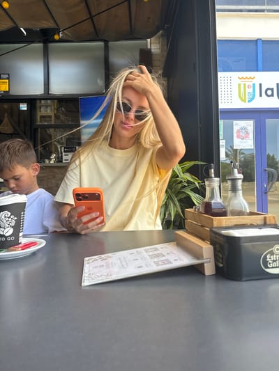 A young woman with blonde hair and sunglasses is holding an orange smartphone and taking a selfie at an outdoor cafe. She is wearing a light yellow t-shirt. A young boy in a white t-shirt is visible in the foreground, seemingly looking down at something on the table.
The setting appears to be a cafe or restaurant with an outdoor seating area. In the background, there is a building with blue and white signage that includes text and a logo. There are also some plants and bottles of what appear to be condiments on a wooden stand. A menu or flyer is placed on the table in front of the woman. The lighting suggests it is daytime, possibly late afternoon, with a mix of natural light and shade. The overall mood seems relaxed and casual.
Visible text includes "la Vila Joiosa" and "LA VILA JOIOSA" on the signage in the background, along with "MOSTAR" and "EXITADA A POLICIA" on a yellow sticker on a wooden structure in the lower left of the image. The coffee cup in the foreground has text that includes "TO GO". The woman's phone case is bright orange.