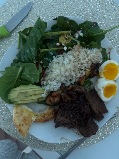 A plate contains a diverse meal. There are two halves of a boiled egg, a mound of white rice, and sliced beef with cooked onions. A spinach salad with feta cheese and sun-dried tomatoes occupies another section, alongside sliced avocado and a piece of cheesy flatbread. A knife with a green handle is visible on the left, and a fork is on the right.