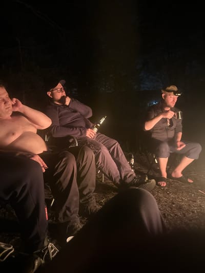 The image shows a group of people in a dark outdoor setting, possibly in the city of Юзуфоўскі сельскі Савет, Belarus. The lighting is poor, with most of the scene shrouded in darkness. Several individuals are visible, some sitting or reclining on what appear to be folding camping chairs. One person is shirtless, and another is wearing a headlamp. People are holding bottles, suggesting they may be drinking. The surrounding environment is unclear, but the presence of grass and vegetation indicates an outdoor location.