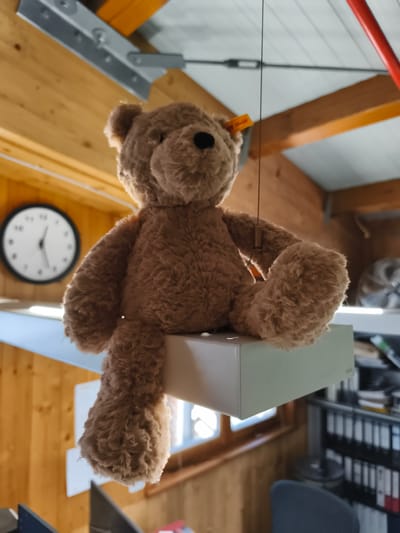 A plush brown teddy bear is suspended by a thin string from the ceiling of a room with wooden walls and ceiling. The bear is positioned in the foreground, with its left leg dangling off the edge of a white shelf. A round clock with a black frame is visible on the wall to the left of the bear. The background shows shelves filled with binders and office supplies, and a partially visible office chair. The lighting suggests an indoor setting, likely an attic or workshop, given the exposed wooden beams and industrial-style metal fixtures on the ceiling. The scene does not provide any specific clues to pinpoint the location within Triberg im Schwarzwald, Germany.