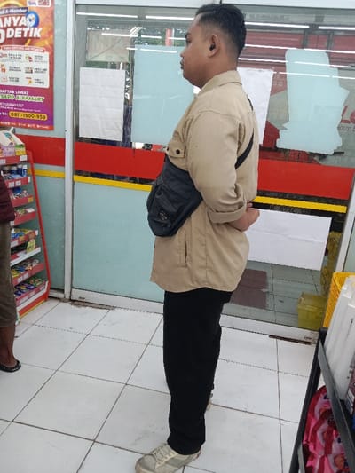 The image shows a man standing in front of a glass storefront, wearing a light brown long-sleeved shirt, dark pants, and a black crossbody bag. He is facing the left side of the image. The glass storefront has a red and yellow stripe across the top and a white sheet on the bottom. To the left of the man, there is a rack of colorful snacks on a white shelf. The floor is tiled in white and the background is blurred. This is the location of Alfamart at Jalan Raya Brangsong - Demak, Kecamatan Brangsong, Kabupaten Demak, Jawa Tengah 59562, Indonesia. The coordinates -6.945, 110.224, suggest the location is in Kecamatan Brangsong, Indonesia.