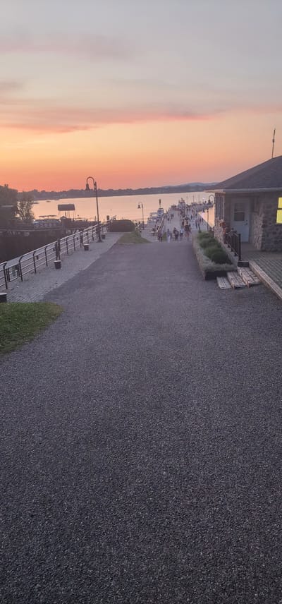 The image depicts a waterfront scene at sunset in Chambly, Canada, likely along the Chambly Canal. In the foreground, a gravel pathway slopes gently upwards towards a boardwalk. A black metal railing lines the left side of the pathway, separating it from a darker, lower area with some green grass. Beyond the railing, a body of water is visible, with several boats docked.
The boardwalk extends into the distance, populated by numerous people walking and gathered. Several lamp posts are spaced along the boardwalk, and a stone building with a lit window is situated on the right side of the frame, close to the foreground. The sky is a gradient of soft pinks and oranges, characteristic of a sunset, with distant hills visible on the horizon. The overall atmosphere is peaceful and recreational.