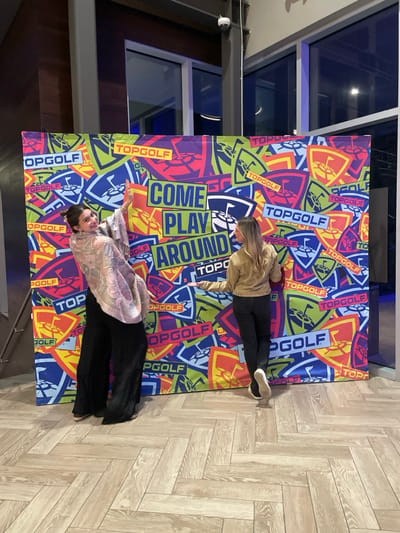 Two women are posing on a "Topgolf" backdrop. One woman has blonde hair and is wearing a tan sweater with black pants and white sneakers. The other woman has dark hair and is wearing a pink and white floral print shirt with black pants. The backdrop is covered in "Topgolf" logos and the phrase "Come Play Around". The floor is light-colored wood in a herringbone pattern.