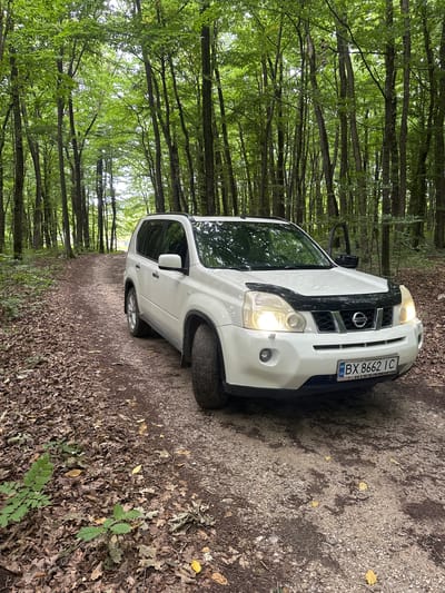 A white Nissan X-Trail SUV is parked on a dirt road in a forest. The car's license plate reads "BX 8662 IC". The road is covered with fallen leaves and is lined by tall trees with green foliage. The sun is shining through the trees, casting dappled light on the scene. The image was taken in the village of Kolubaivtsi, Ukraine.