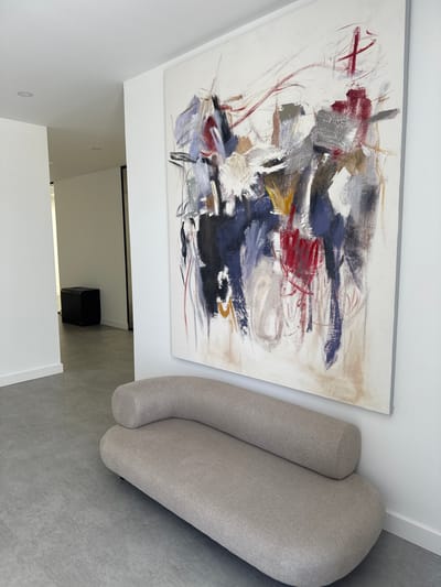 A contemporary interior shot from an elevated perspective shows an abstract painting on a white wall, adjacent to a light gray rounded couch. The painting is composed of red, blue, black, white, and gray brushstrokes. Below the painting, a doorway with a black frame leads to a space with a black cube-shaped object. The floor is gray, and the walls are white. The location is possibly Torres Vedras, Portugal, though this is not explicitly verifiable from the image. No people are visible.
