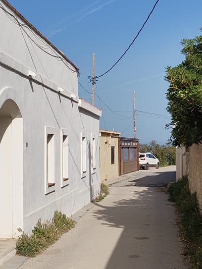 A narrow asphalt road, flanked by buildings, stretches into the distance in Rotes, Spain. On the left, a white building with a light gray façade, marked by three window openings, rises. The bases of its walls are lined with small patches of green vegetation. To the right of this building is a tan structure with an addition of dark brown siding and windows. In the background, a white SUV is parked on the road. Power lines stretch across the sky, connecting wooden utility poles. The sky is clear and blue. A stone wall overgrown with greenery borders the right side of the road, partially obscuring the view. Shadow patterns from unseen elements are cast across the road.