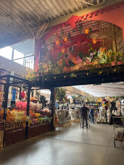 This indoor shot depicts a section of Famiflora in Mouscron, Belgium. The upper left portion of the image features a highly decorated, red-themed ceiling display adorned with tropical foliage, cage-like structures, and decorative lighting. Below this, a display of artificial flowers and lollipops on stands occupies the lower portion. The right side shows shelves filled with garden ornaments and statuary, with a couple of shoppers browsing. The floor is a light grey concrete. Overhead, the ceiling structure is visible, indicating a large retail space.